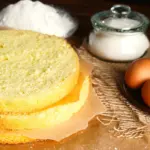 Frisch gebackener Biskuitboden auf einem Kuchengitter, mit Eierschale, Mehl und Schneebesen im Hintergrund – perfekt für Torten und einfache Kuchen.